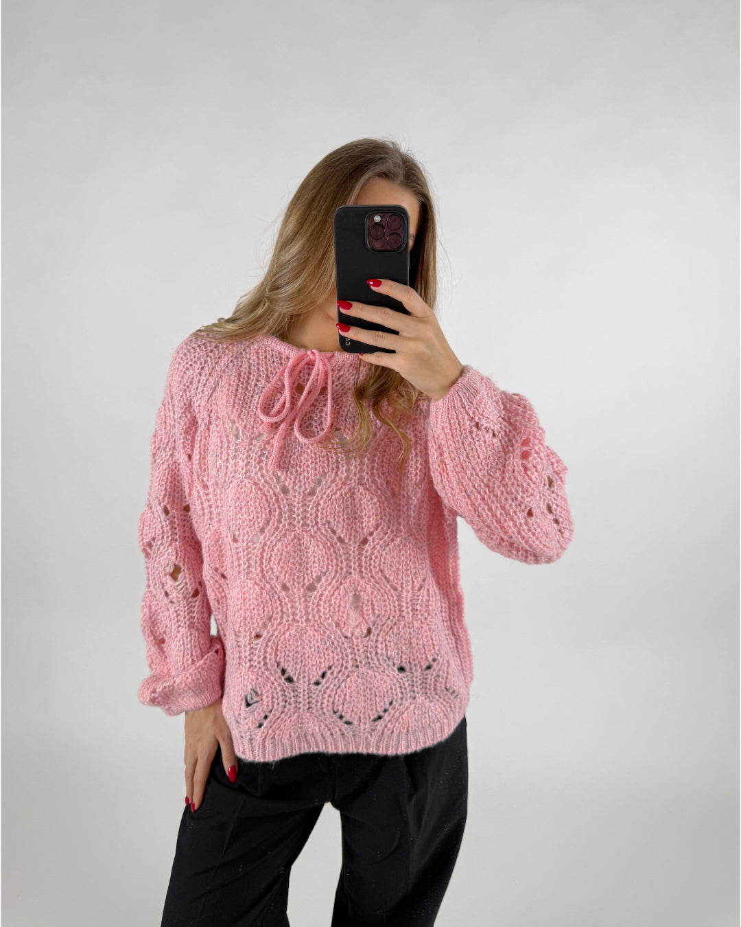 En person iført en lyserød strikket sweater tager en selfie med en mobiltelefon. Personen står i et minimalistisk, lyst rum og har sort tøj på.