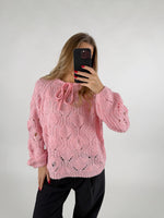 En person iført en lyserød strikket sweater tager en selfie med en mobiltelefon. Personen står i et minimalistisk, lyst rum og har sort tøj på.