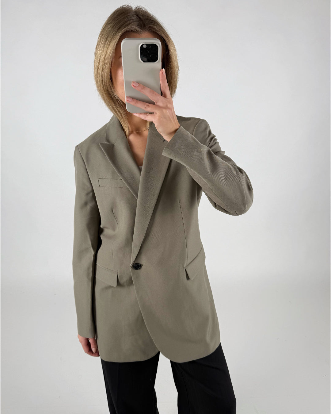 En person iført en grå blazer tager et spejlbillede med en smartphone, der dækker ansigtet, mod en enkel, lys baggrund.