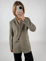 En person iført en grå blazer tager et spejlbillede med en smartphone, der dækker ansigtet, mod en enkel, lys baggrund.