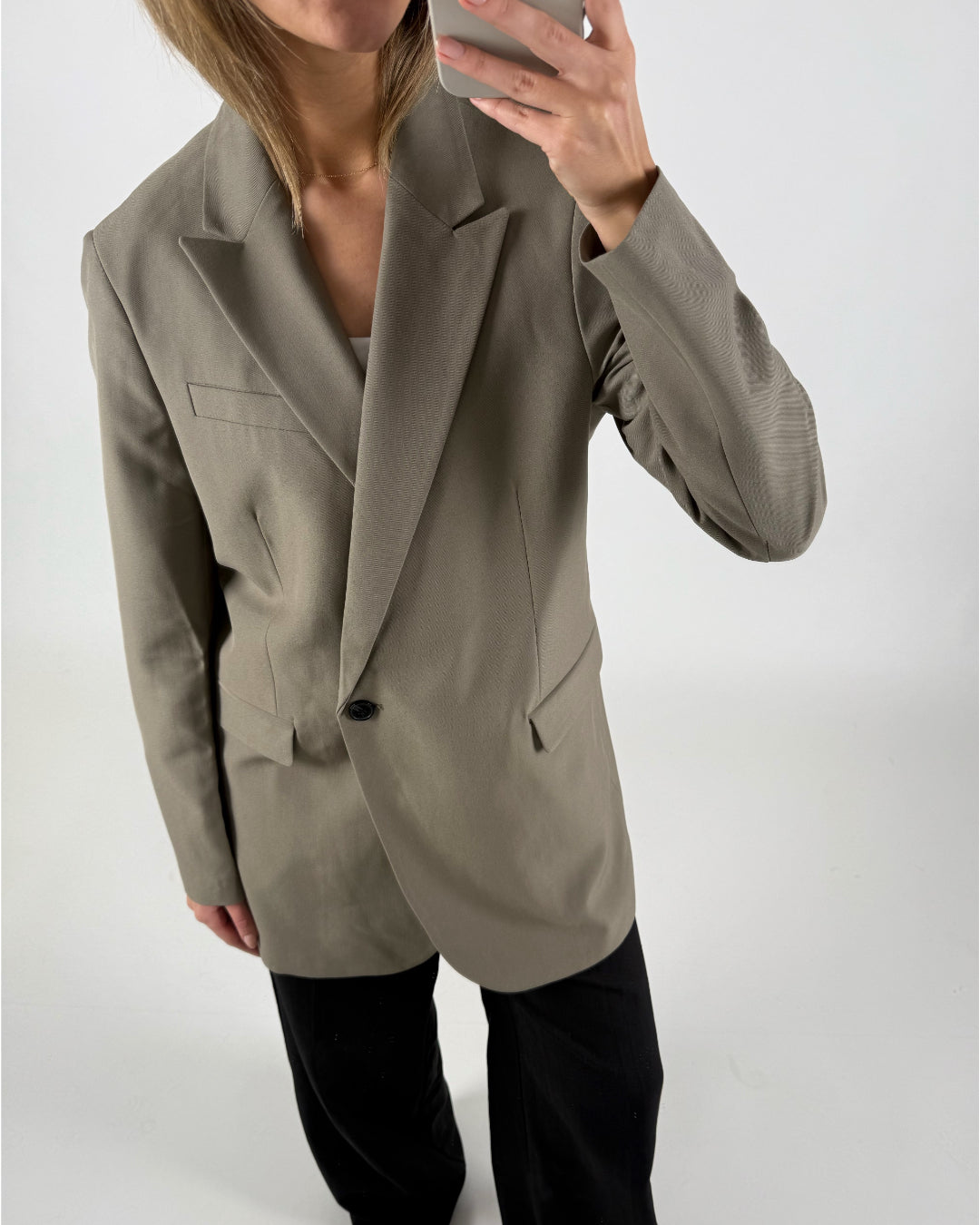 En person iført en beige blazer og sorte bukser tager et spejlbillede med en smartphone i et minimalistisk rum.