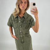 Kvinde iført en olivengrøn denimkjole tager en selfie med sin smartphone i et neutralt, lyst rum. Hun poserer med hovedet let skråt og læberne fremad.