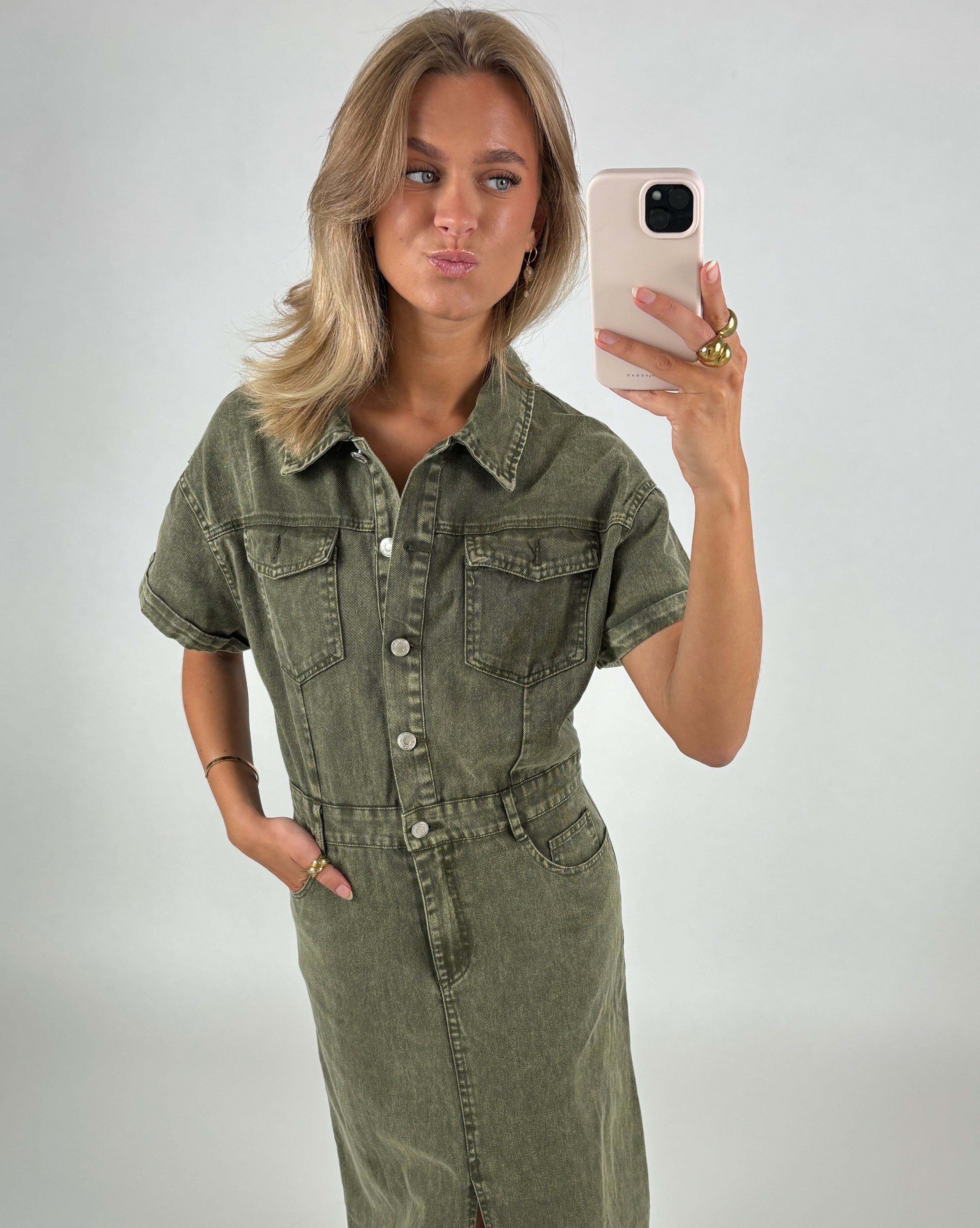 Kvinde iført en olivengrøn denimkjole tager en selfie med sin smartphone i et neutralt, lyst rum. Hun poserer med hovedet let skråt og læberne fremad.