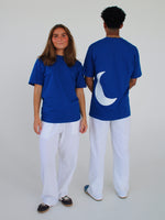 To personer i blå T-shirts og hvide bukser står foran en neutral baggrund; den ene T-shirt har en hvid halvmåne form på ryggen.
