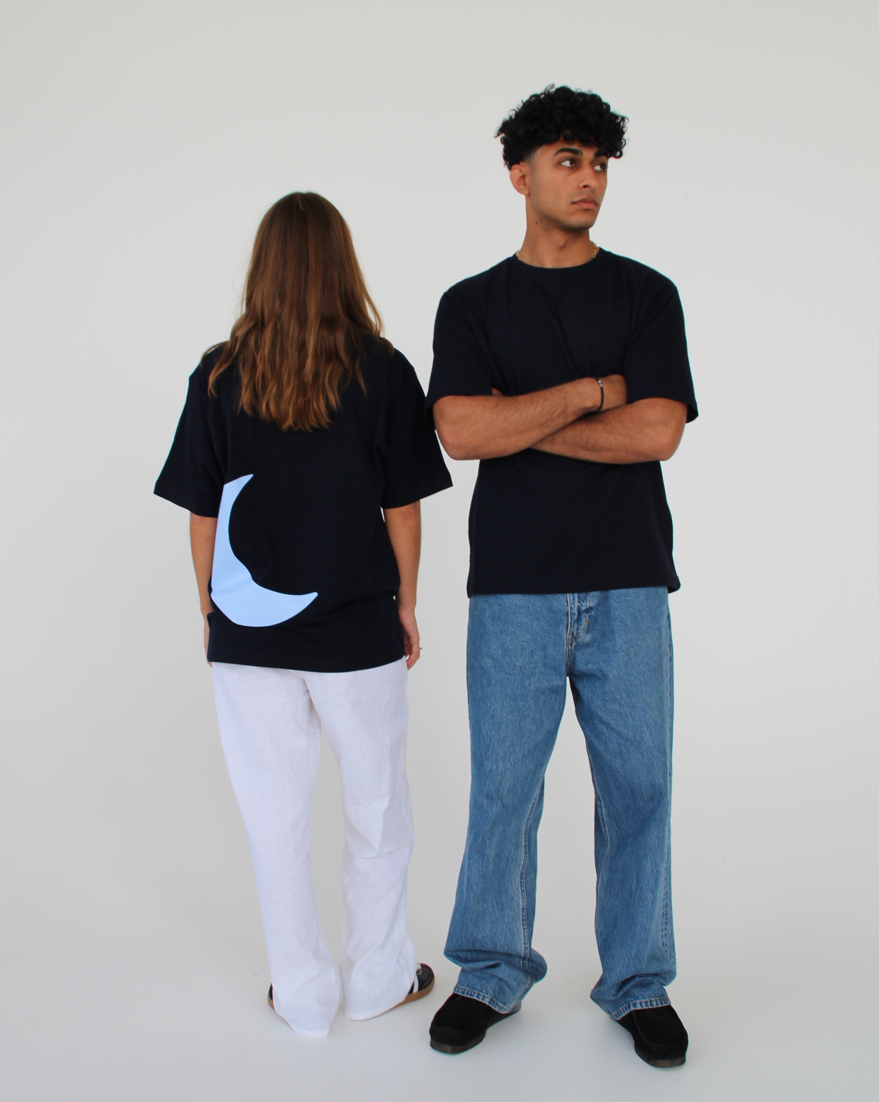 To personer iført sorte T-shirts. Den ene står med ryggen til og har et blåt halvmånemotiv. Den anden står med krydsede arme og kigger til siden i et hvidt rum.