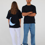 To personer står i et lyst rum. Den ene bærer en sort t-shirt med en blå halvmåne og hvide bukser, vendt bagud. Den anden har en sort t-shirt, krydsede arme og jeans.
