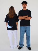 To personer står i et lyst rum. Den ene bærer en sort t-shirt med en blå halvmåne og hvide bukser, vendt bagud. Den anden har en sort t-shirt, krydsede arme og jeans.