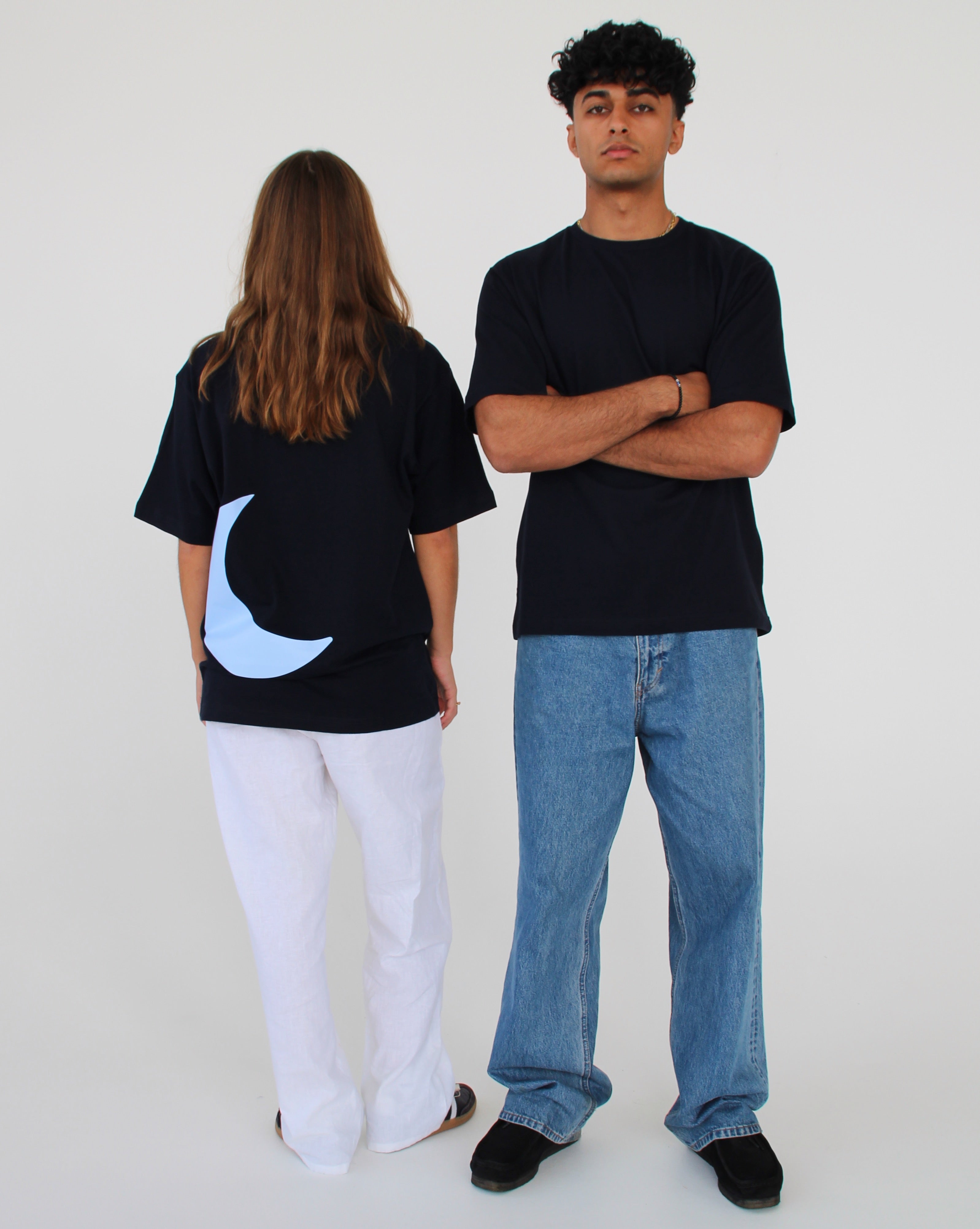 To personer står i et lyst rum. Den ene bærer en sort t-shirt med en blå halvmåne og hvide bukser, vendt bagud. Den anden har en sort t-shirt, krydsede arme og jeans.