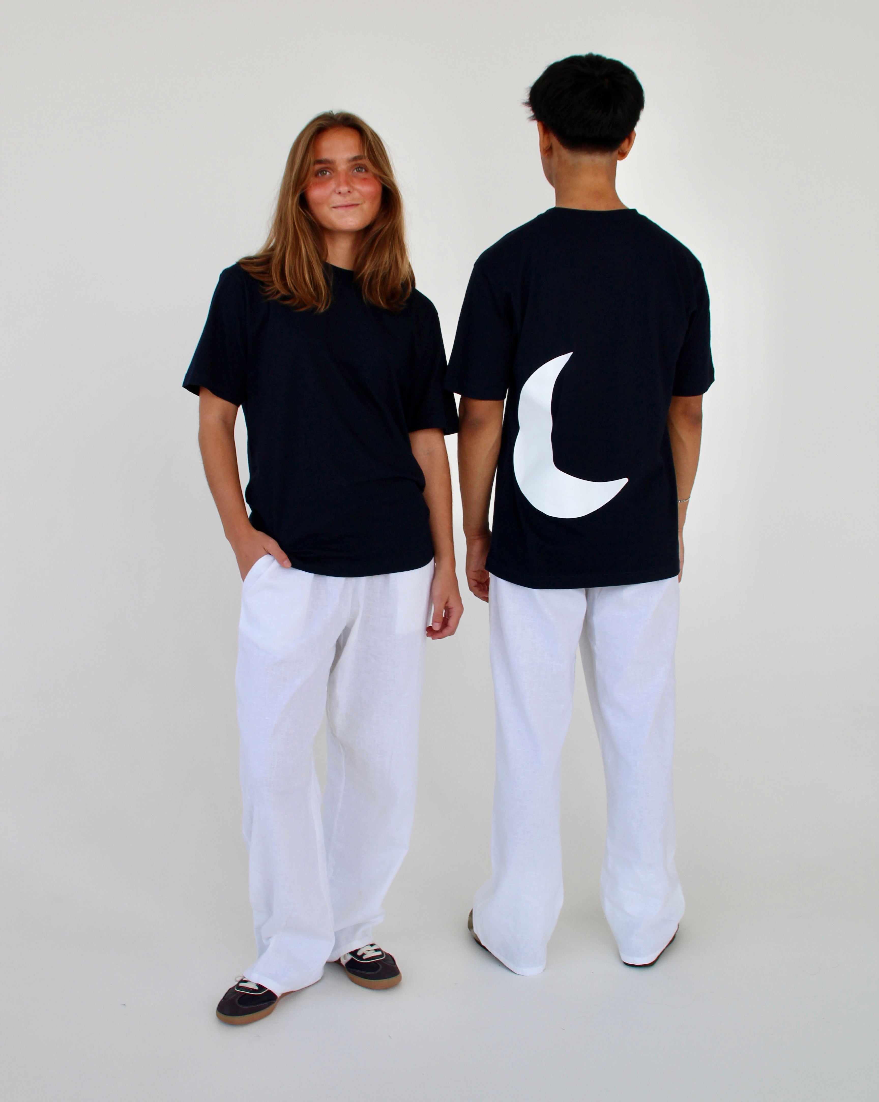 To personer i sorte t-shirts og hvide bukser står sammen; den ene vender mod kameraet, den anden med ryggen til. T-shirten har en stor hvid halvmåne bagpå.