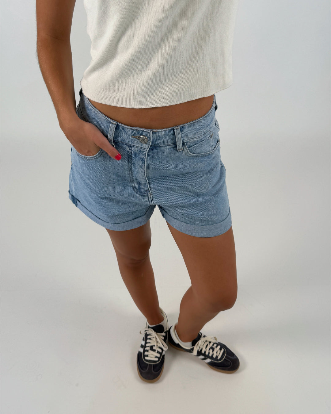 En person iført lyseblå denimshorts og hvide sneakers står med hænderne i lommerne på en lys, ensfarvet baggrund.