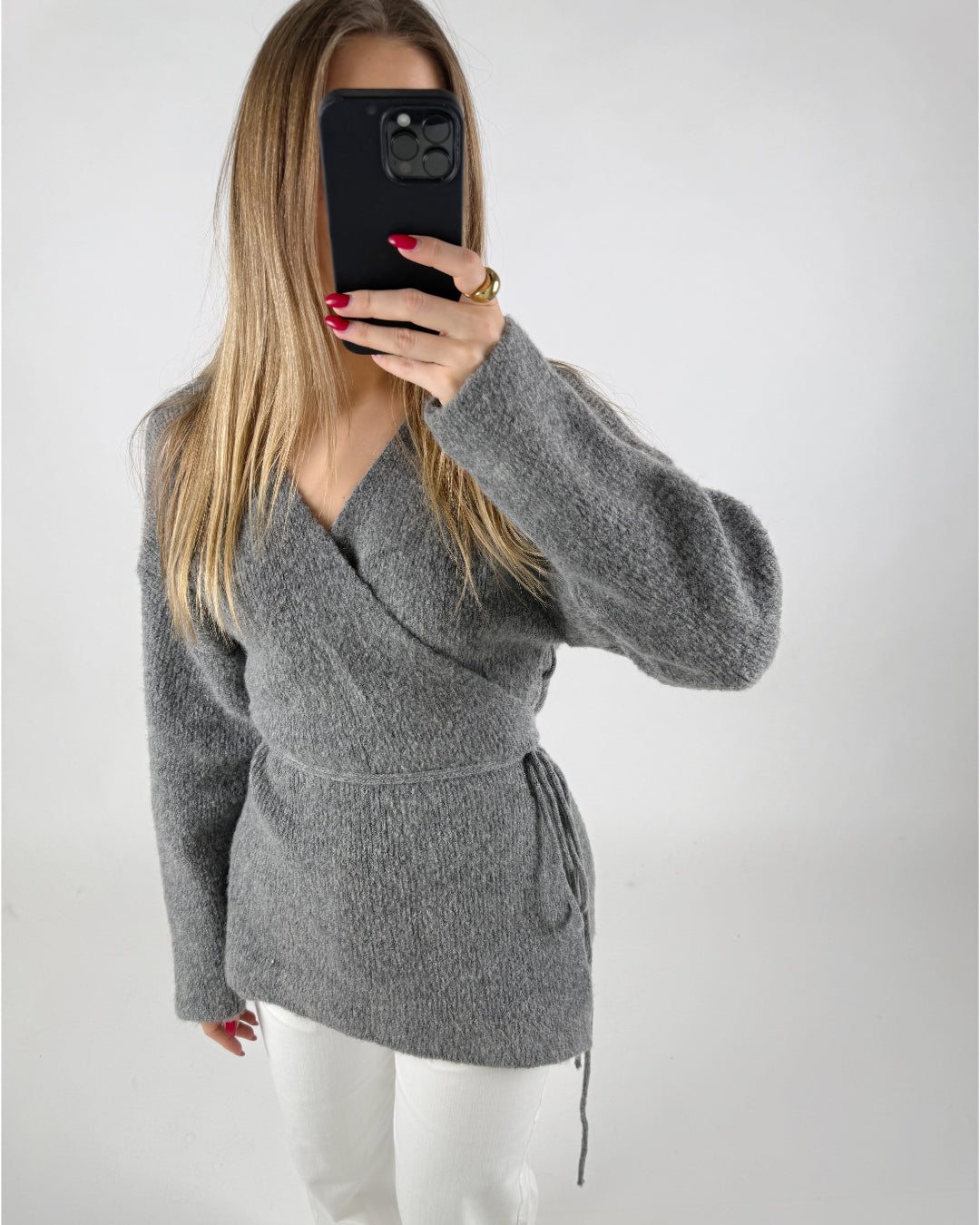 En person iført en grå sweater tager et spejlbillede med en sort mobiltelefon; personen har langt, lyst hår og rødmalede negle, stående i et enkelt, hvidt rum.