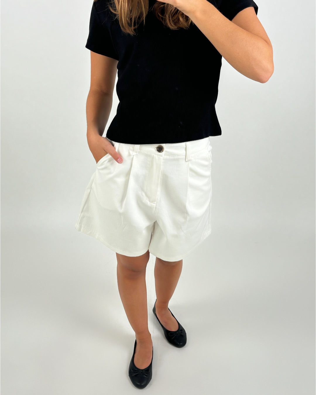En person iført sort t-shirt og hvide shorts står med en hånd i lommen. Personen bærer sorte flade sko, og baggrunden er ensfarvet lys.