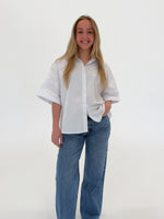 En person i en hvid skjorte står og smiler, med hånden i lommen på jeans. Baggrunden er ensfarvet og neutral, uden tekst.