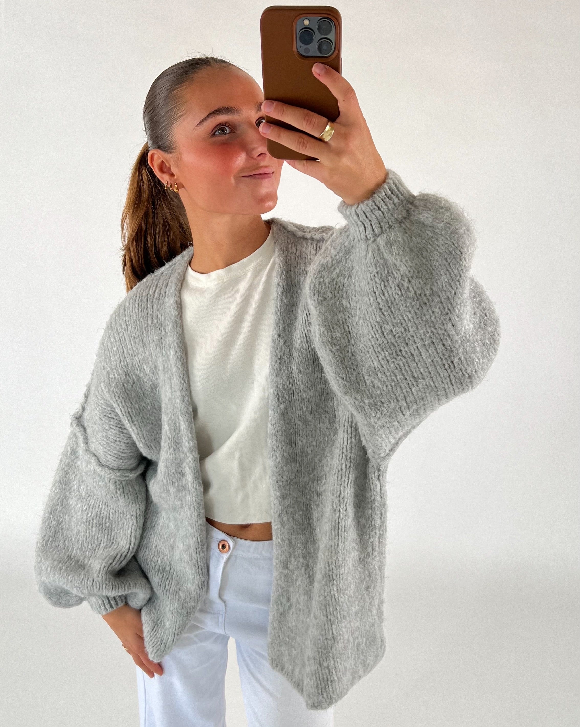 En person tager en selfie iført en grå striktrøje og hvid bluse. Personen holder en telefon med brun cover i et lyst rum.