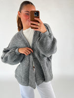 En person iført en grå, strikket cardigan tager en selfie med en telefon i en brun etui. Personen bærer en hvid t-shirt og hvide bukser i et lys rum.