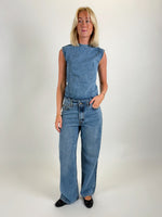 En person iført en ærmeløs denim top og jeans står oprejst i et lyst rum, ser fremad. Vedkommende har lyst hår og sorte flade sko.