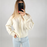 En person i en hvid sweater tager et billede med en mobiltelefon, der dækker ansigtet. De har lyseblå jeans på og står i et minimalistisk rum.