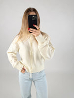 En person i en hvid sweater tager et billede med en mobiltelefon, der dækker ansigtet. De har lyseblå jeans på og står i et minimalistisk rum.