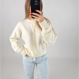 En person iført hvid cardigan og lyseblå jeans tager et spejlbillede med en sort telefon. Personen står i et neutralt belyst rum.