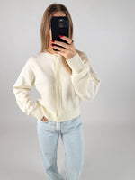 En person iført hvid cardigan og lyseblå jeans tager et spejlbillede med en sort telefon. Personen står i et neutralt belyst rum.