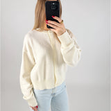 En person iført en cremefarvet cardigan tager et spejlbillede med en mobiltelefon. Personen har rødlakerede negle og en stor ring. Baggrunden er hvid og minimalistisk.