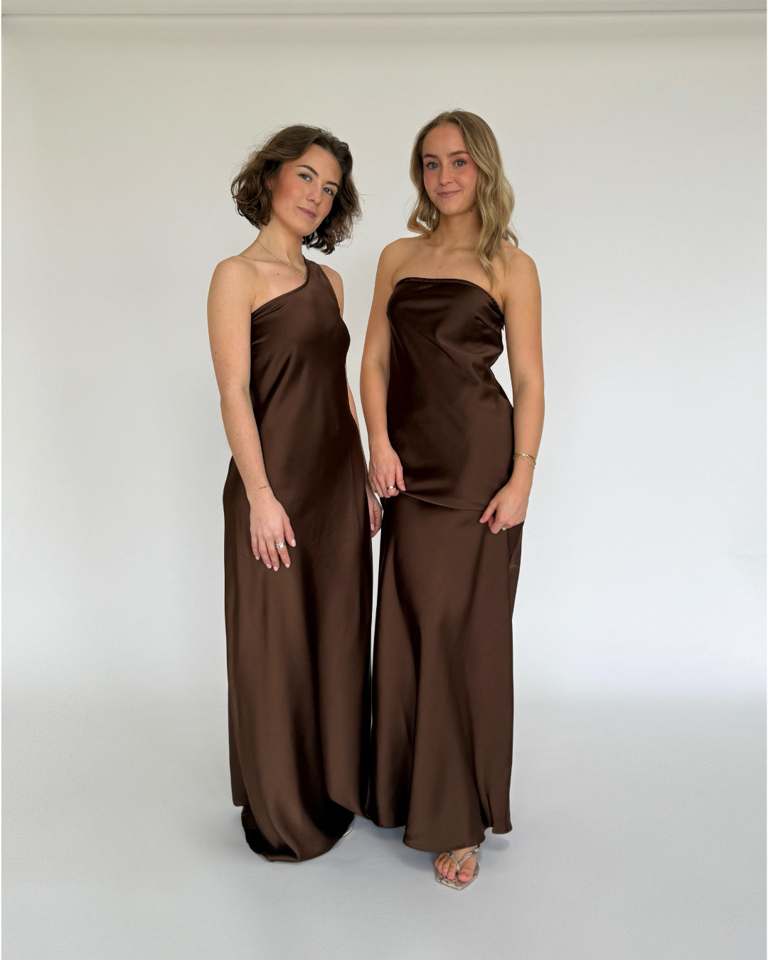 Modelbillede af to piger i brune NAT kjoler, hvor den ene viser en stropløs kjole og den anden viser en one shoulder kjole. Set forfra. Fra BySofieSønderby