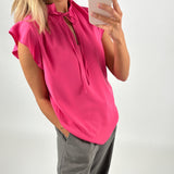En person tager et spejlfoto iført en pink bluse og grå bukser i et lyst rum. Personen holder en mobiltelefon.