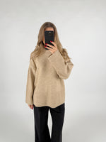 En person iført en beige sweater tager et selfie med en mobiltelefon, mens de står foran en lys baggrund. De har løst, bølget hår og sorte bukser.