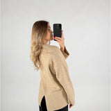 En kvinde i en beige sweater tager et selfie med sin telefon mod en lys baggrund. Hun har langt, bølget hår og bærer sorte bukser. Ingen tekst er til stede.