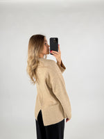En kvinde i en beige sweater tager et selfie med sin telefon mod en lys baggrund. Hun har langt, bølget hår og bærer sorte bukser. Ingen tekst er til stede.
