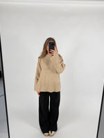 En person står i et lyst rum og tager et spejlbillede iført en beige sweater og sorte bukser. Telefonen dækker ansigtet.