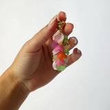 Hånden holder et farverigt nøgleringvedhæng med flere lag af blomstformede plaststykker i pink, orange, grøn og gul toner på en hvid baggrund.