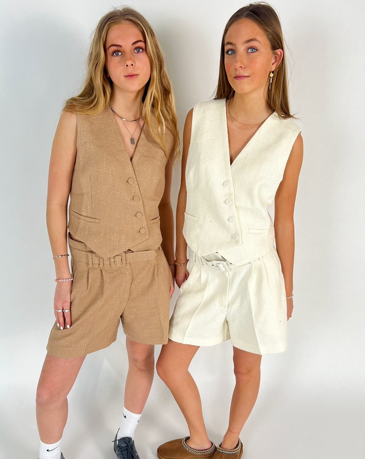 To kvinder iført matchende vest-og-shorts-sæt, den ene i beige, den anden i hvid, poserer sammen mod en hvid baggrund, iført afslappet fodtøj og smykker.