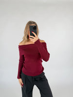 En person iført en mørkerød off-shoulder trøje tager et spejlbillede med en sort telefon i et minimalistisk rum med lysegrå baggrund.