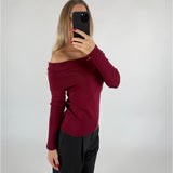 En person tager en selfie med en smartphone, iført en off-shoulder bordeaux trøje og sorte bukser, i et enkelt, lyst rum.