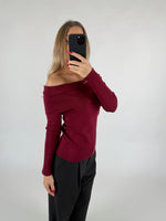 En person tager en selfie med en smartphone, iført en off-shoulder bordeaux trøje og sorte bukser, i et enkelt, lyst rum.