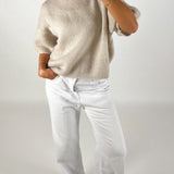 En person iført en løs, beige sweater putter hånden i lommen på hvide bukser. Personen står oprejst i et neutralt, lys rum.