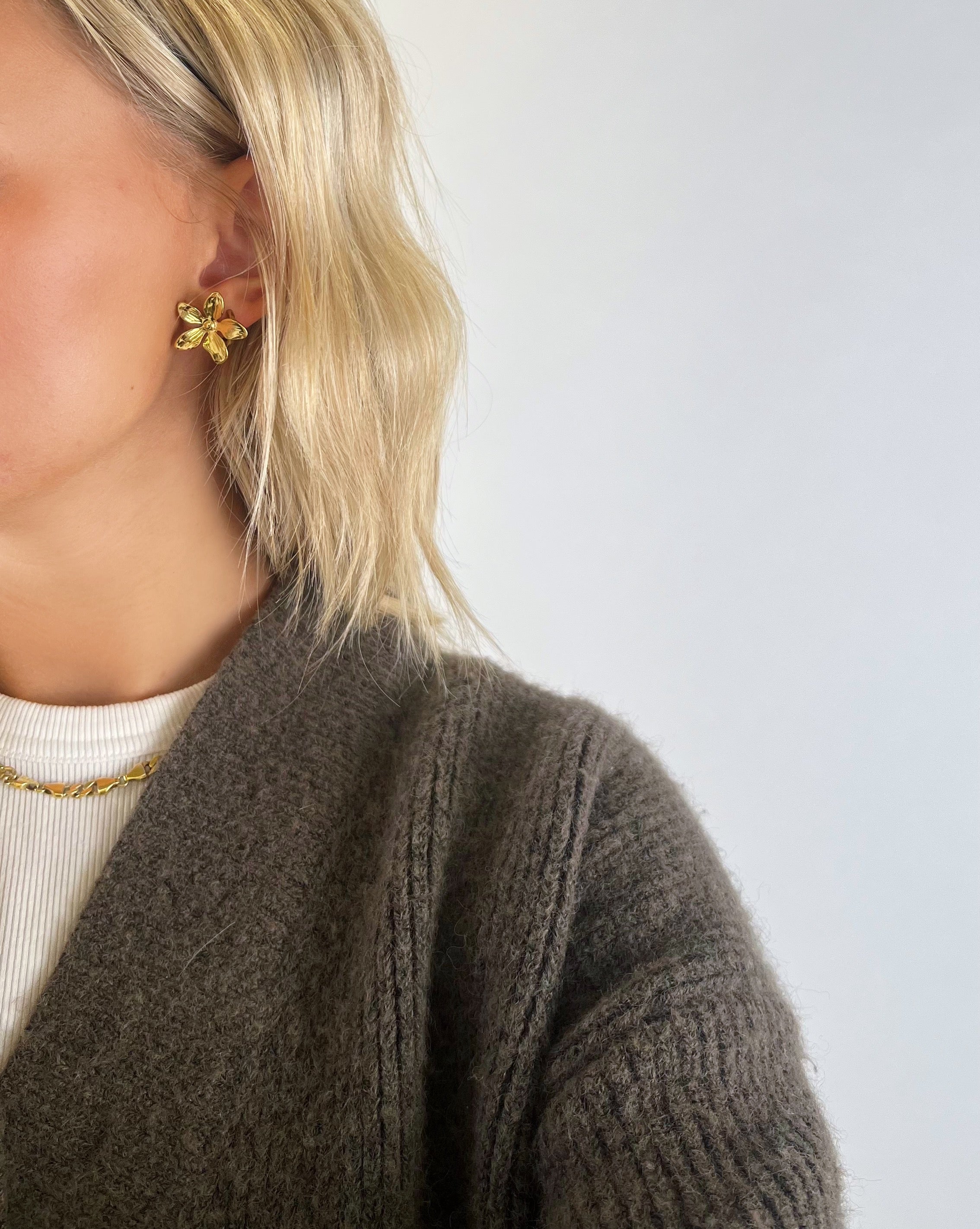 En person med lyst hår bærer en guldfarvet blomsterørering. Personen er iført en brun striktrøje og en hvid bluse med halskæde, mod en neutral baggrund.