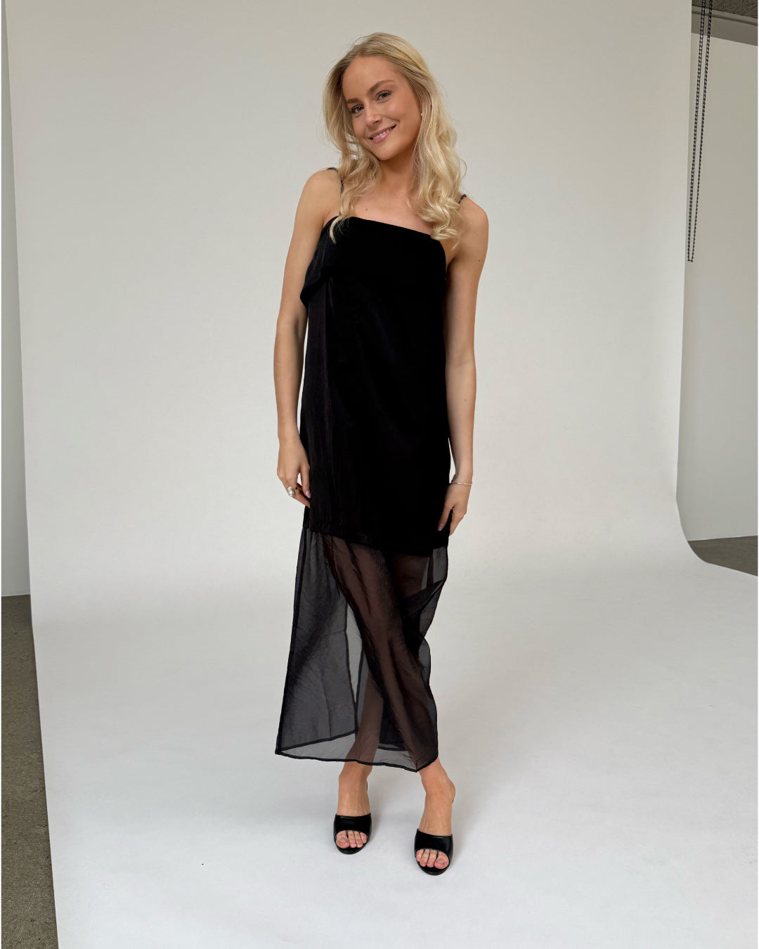 Kvinde i sort kjole står afslappet på en hvid baggrund med let smil, omgivet af fotostudieudstyr.