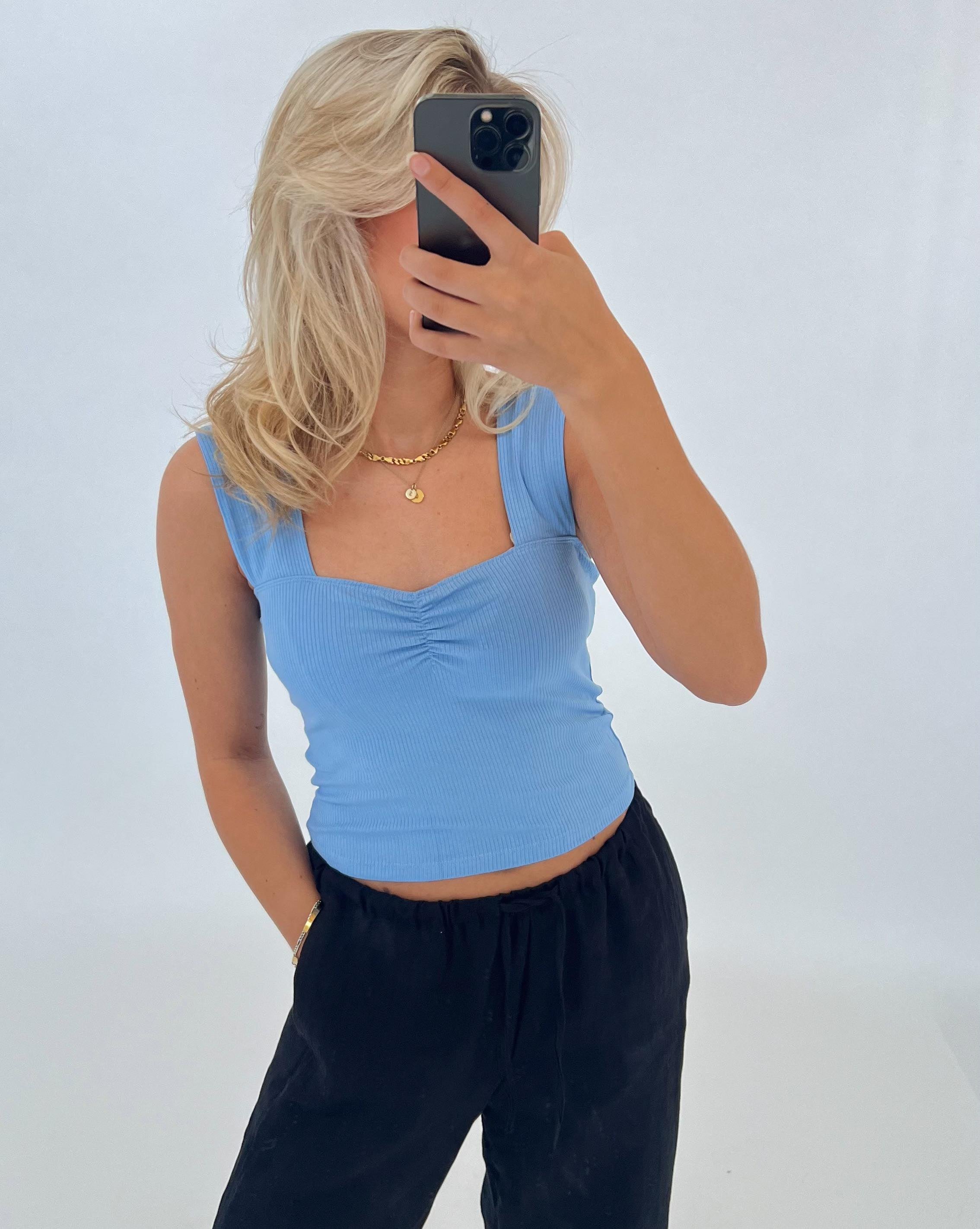 Person tager et spejlbillede med en smartphone. De har en blå top, sort bukser og gyldent smykke på. Baggrunden er helt hvid, hvilket skaber en minimalistisk stil.