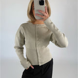 En person i en lys beige cardigan holder en smartphone op foran ansigtet. Personen står i et rum med en hvid baggrund og har en hånd i lommen på sine sorte bukser.