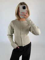 En person i en lys beige cardigan holder en smartphone op foran ansigtet. Personen står i et rum med en hvid baggrund og har en hånd i lommen på sine sorte bukser.