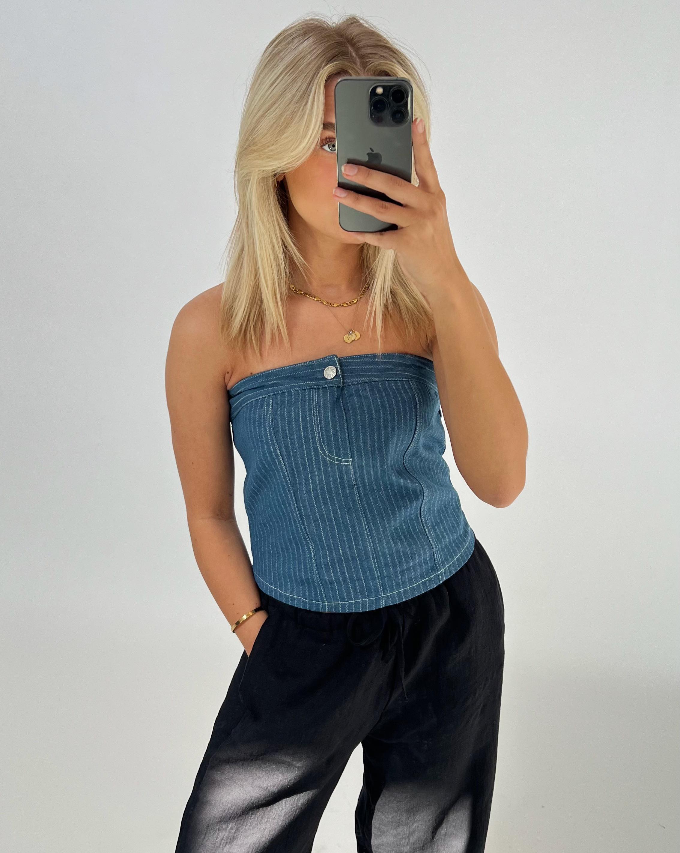 En person tager en selfie med en smartphone, iført en blå stropløs top og sorte bukser, i et enkelt, lyst rum.