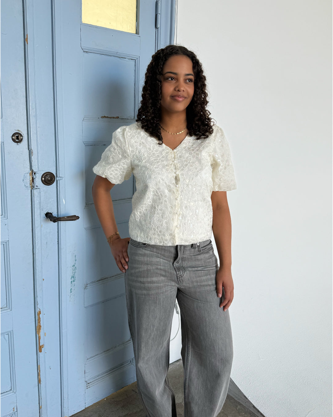 En person står foran en lyseblå dør iført en hvid bluse og grå jeans, smilende let. Baggrunden er en hvid væg, hvilket skaber en rolig atmosfære.