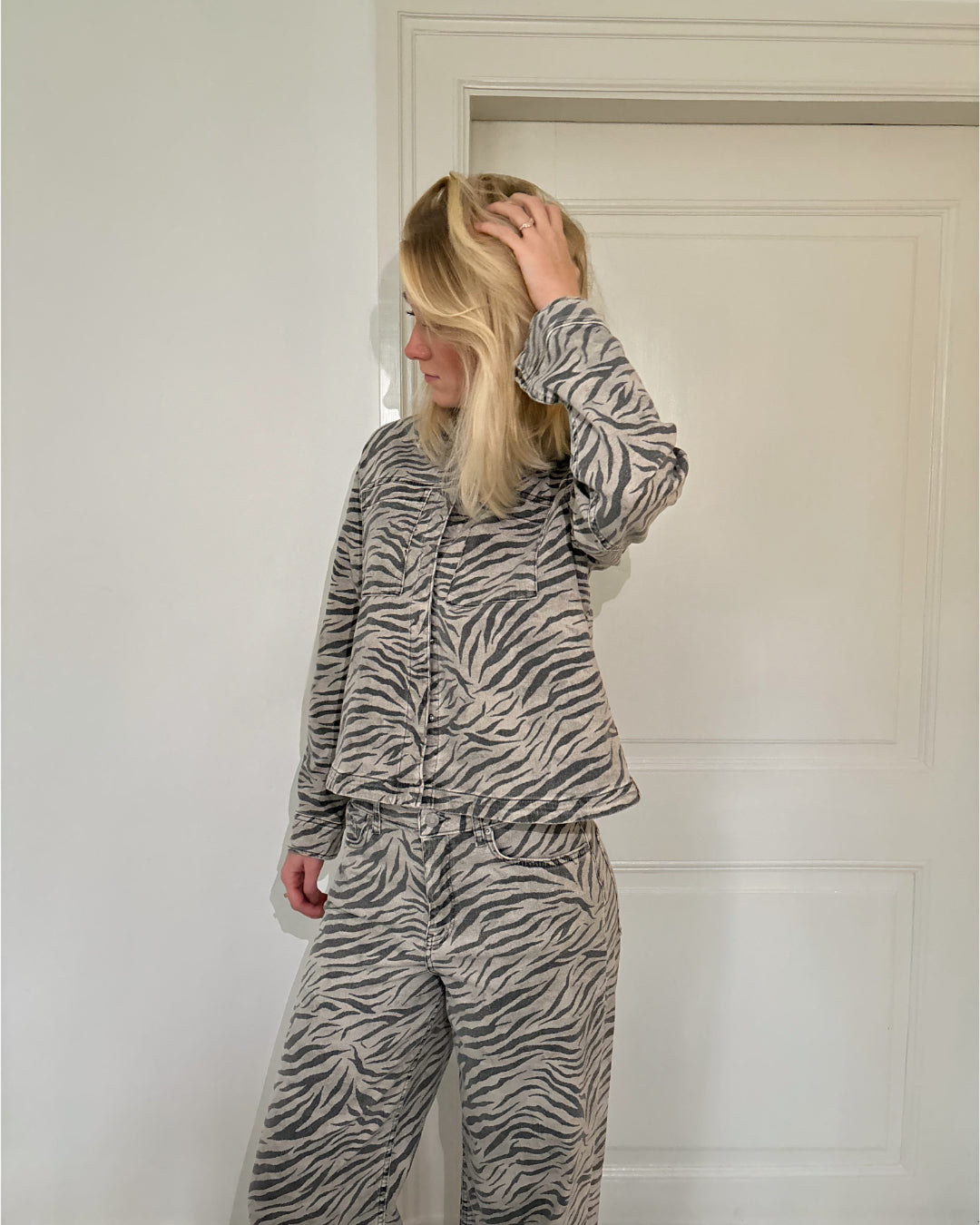 En person iført et zebra-mønstret sæt tøj står med hånden i håret foran en hvid dør i et minimalistisk rum.
