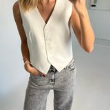 En person med lyseblondt hår bærer en hvid vest og stenvasket jeans, mens de tager en selfie i et minimalistisk rum. Personen har hånden i lommen og bærer et armbånd.