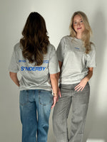 To kvinder iført grå T-shirts står ved siden af hinanden i et neutralt rum. Den ene vender ryggen til, mens den anden ser fremad. T-shirt teksten lyder: "S*NDERBY".