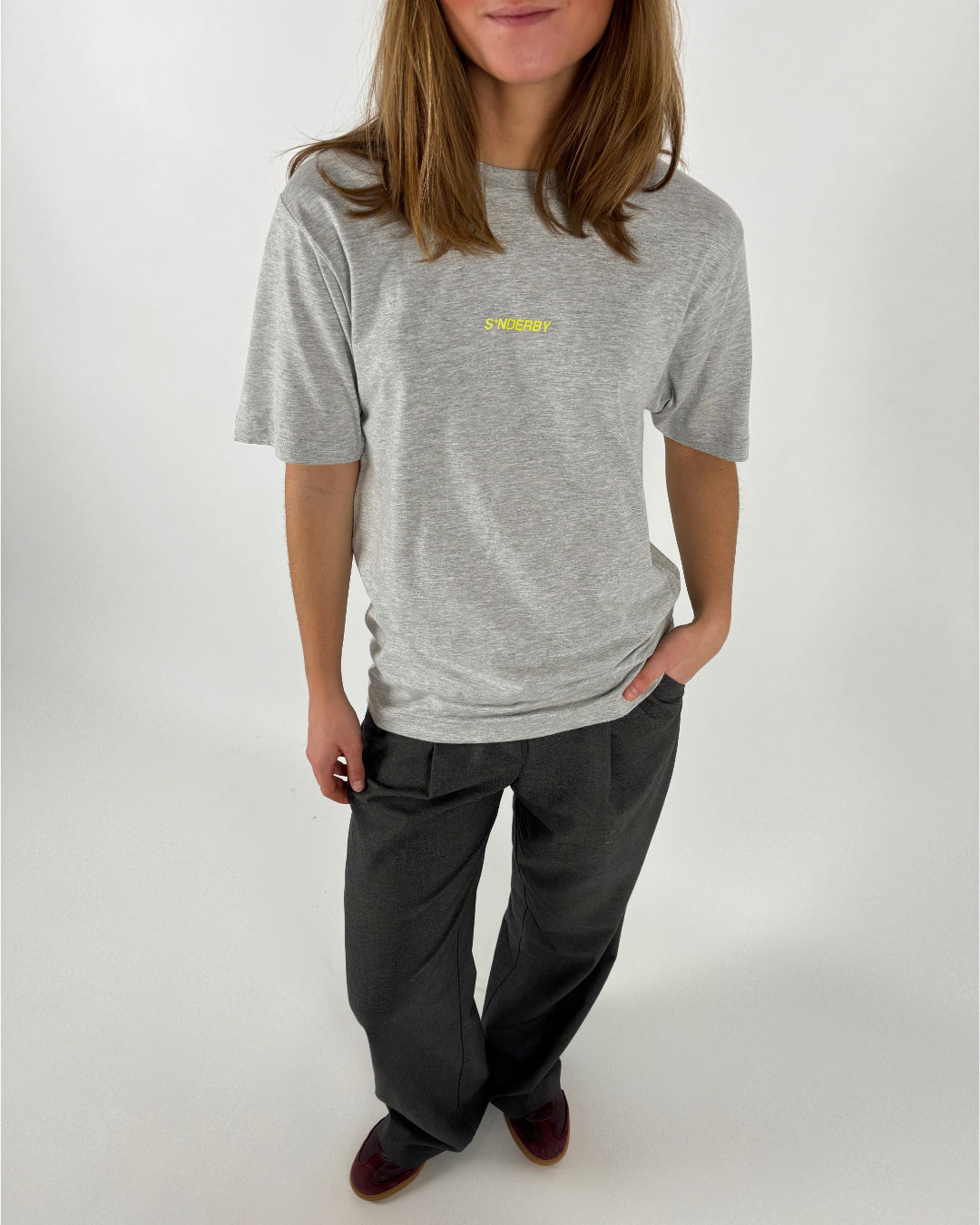 En person står oprejst iført en grå T-shirt med teksten "SØNDERBY" og mørke bukser. Hånden er i lommen, og omgivelserne er enkle og lyse.