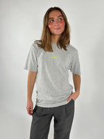 En person iført en grå T-shirt med teksten "S'NOEBY" står afslappet med hånden i lommen på et par mørke bukser, mod en lys baggrund.