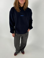 En person iført en sort sweatshirt med teksten "SØNDERBY" står i hvilestilling; bukselommerne hviler i lysegrå bukser med brune sko på et hvidt baggrund.